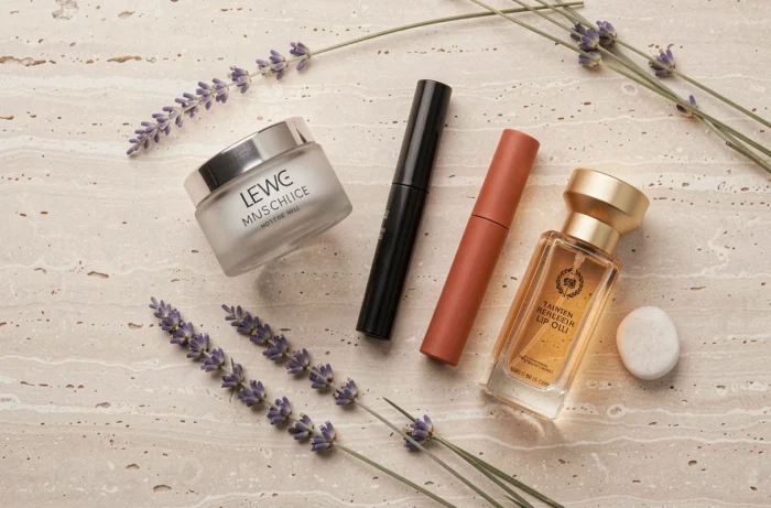 Flat lay de produits de beauté indispensables sur pierre travertine avec mascara, baume lèvres, blush terracotta et parfum